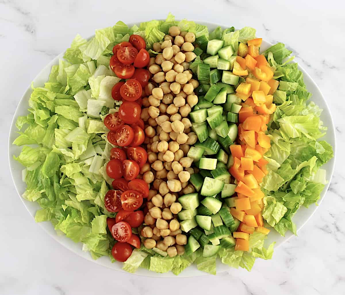 chopped lettuce in white oval platter with chickpeas and chopped cucumber, orange peppers and halved tomatoes down the center