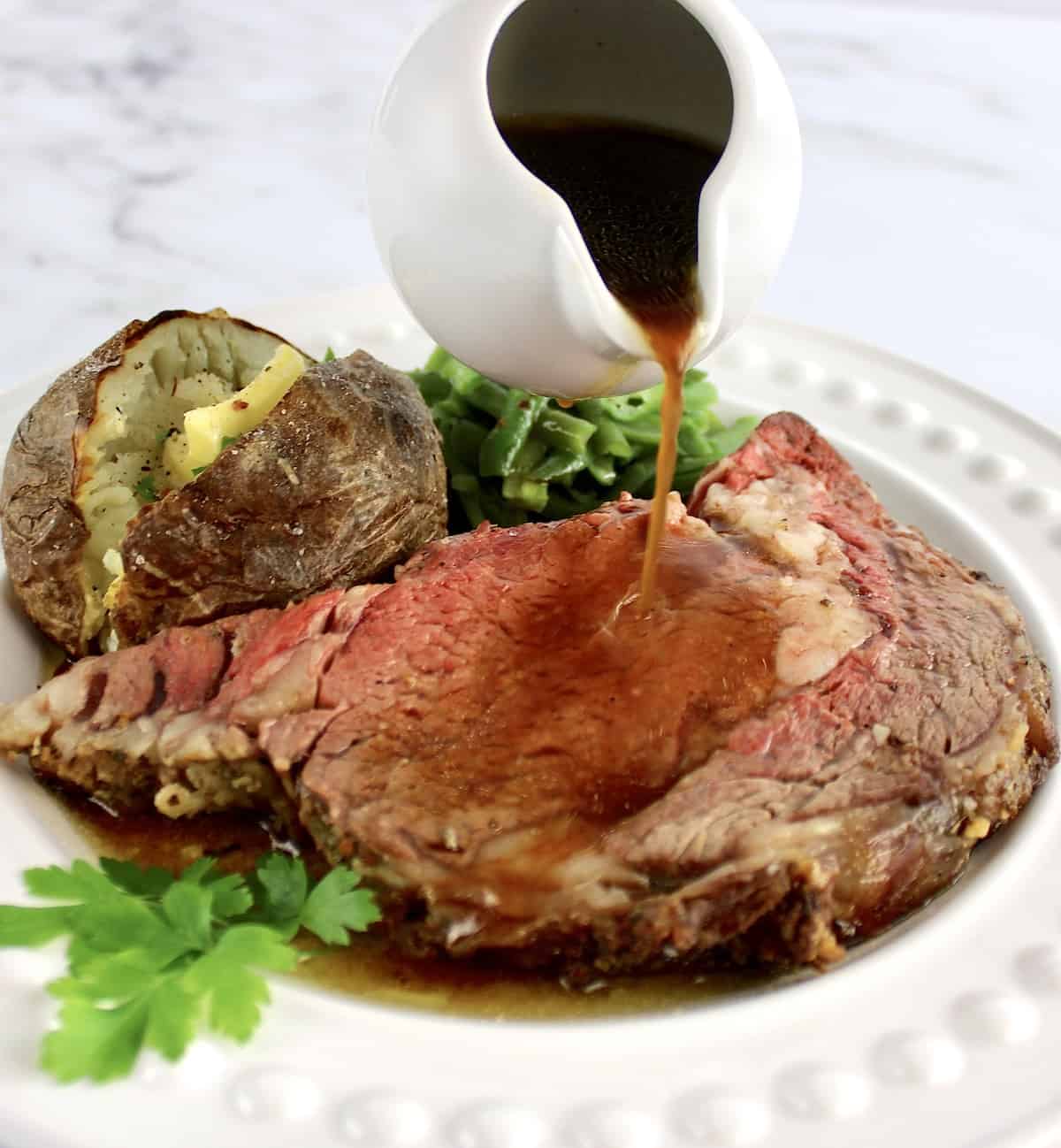 Prime Rib on white plate with baked potato and green beans au jus being poured over steak
