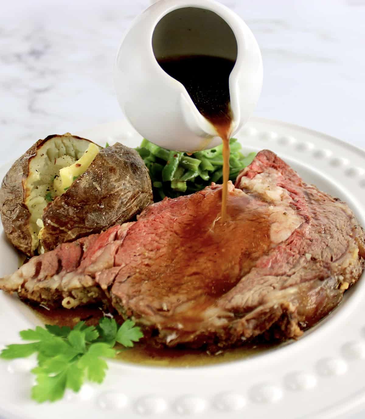 Prime Rib on white plate with baked potato and green beans au jus being poured over steak