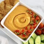 Roasted Red Pepper Hummus in white square bowl with pita chips, cucumbers and tomatoes on side