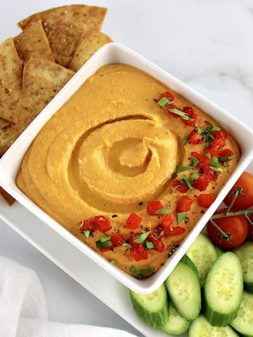Roasted Red Pepper Hummus in white square bowl with pita chips, cucumbers and tomatoes on side