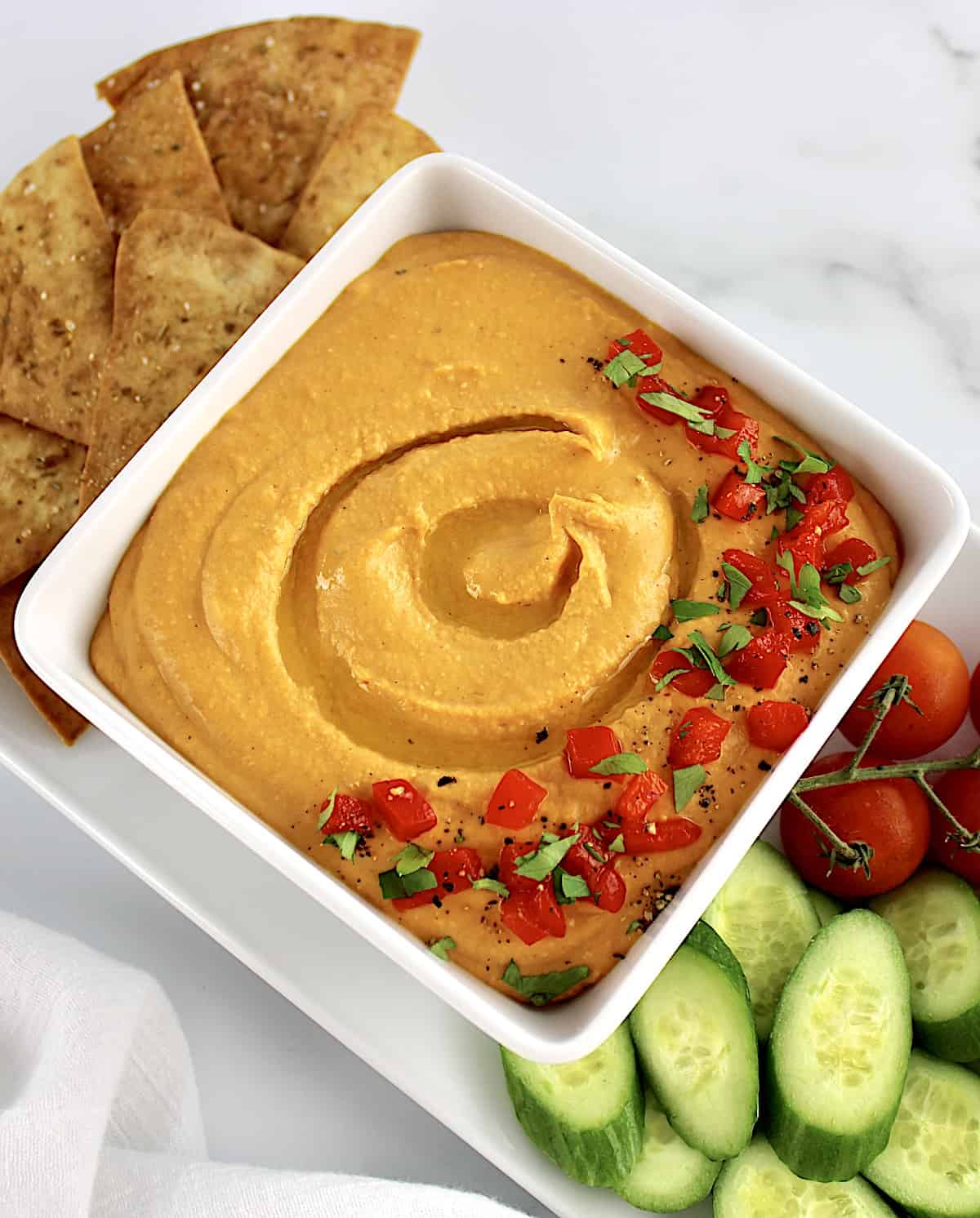 Roasted Red Pepper Hummus in white square bowl with pita chips, cucumbers and tomatoes on side