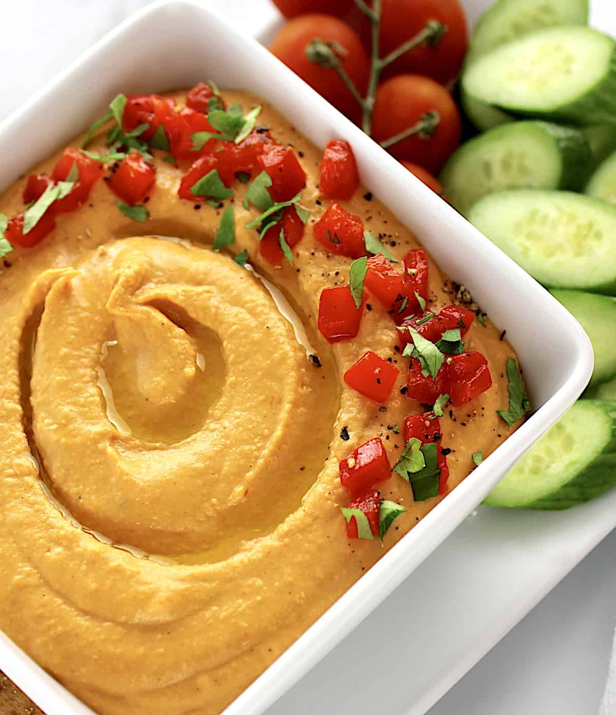 closeup of Roasted Red Pepper Hummus in white square bowl with cucumber slices and cherry tomatoes in background