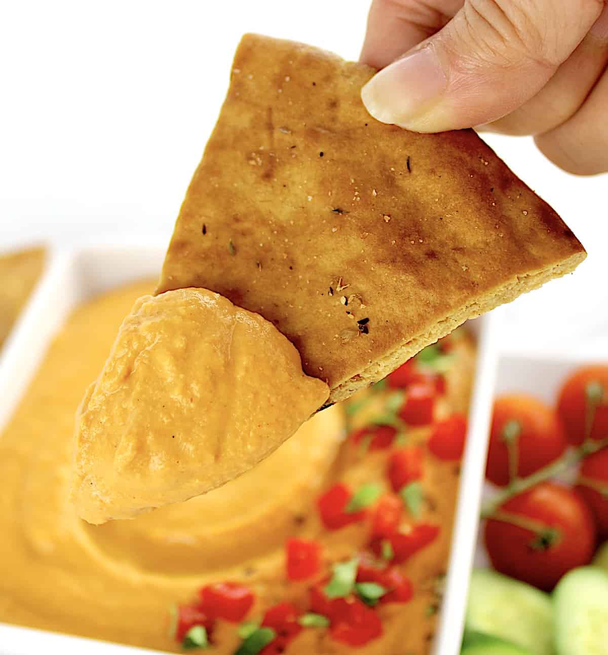holding up pita chip dipped in Roasted Red Pepper Hummus in white bowl