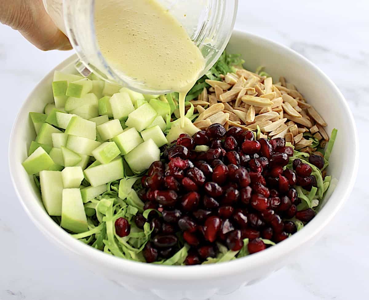 Shredded Brussels Sprouts, pomegranate seeds, slivered almonds, diced green apples and chopped mint in white bowl with dressing being poured on top