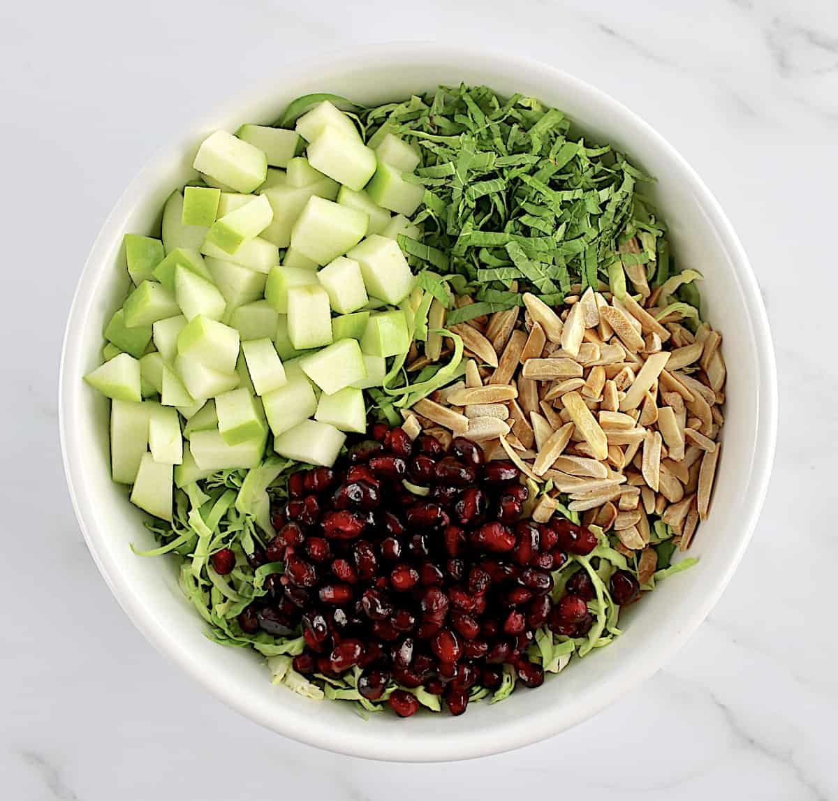 Shredded Brussels Sprouts, pomegranate seeds, slivered almonds, diced green apples and chopped mint in white bowl