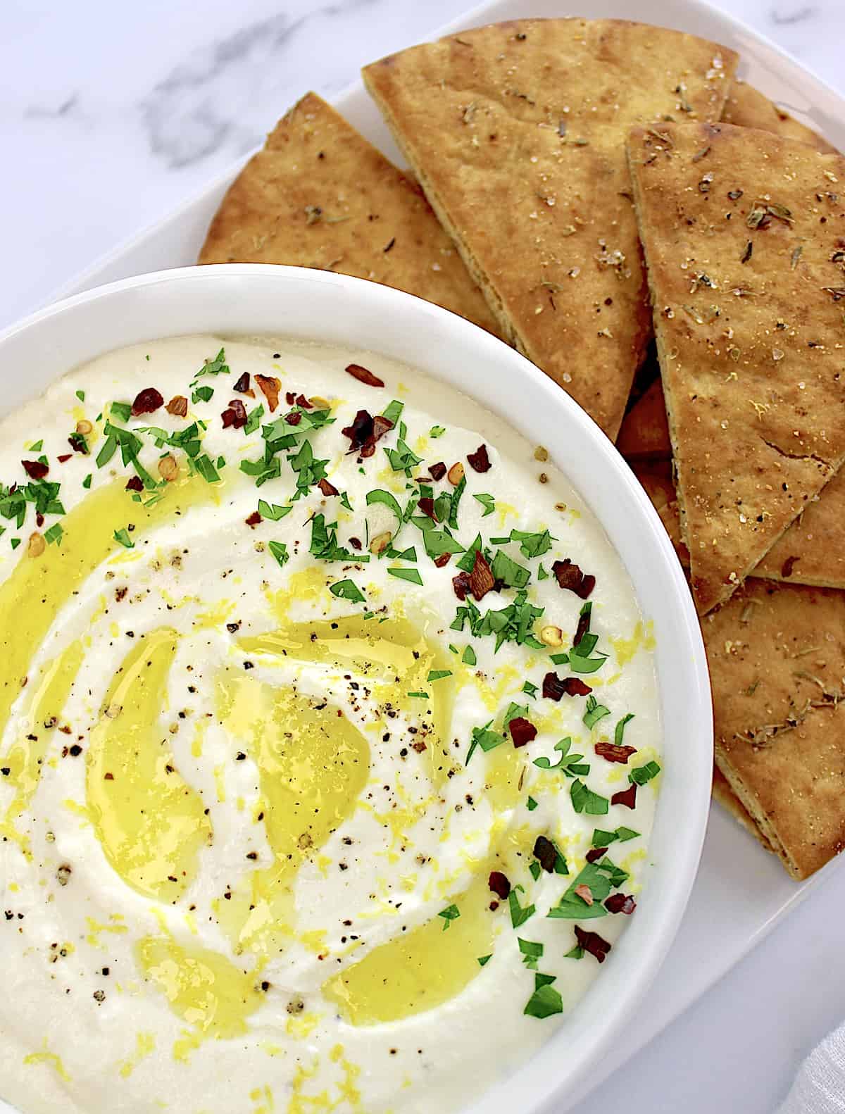 Whipped Feta Dip in white bowl with drizzle of olive oil and herbs, pita chips on side