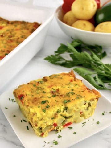 closeup of Sausage and Potatoes Breakfast Casserole slice on white plate with casserole and parsley in background