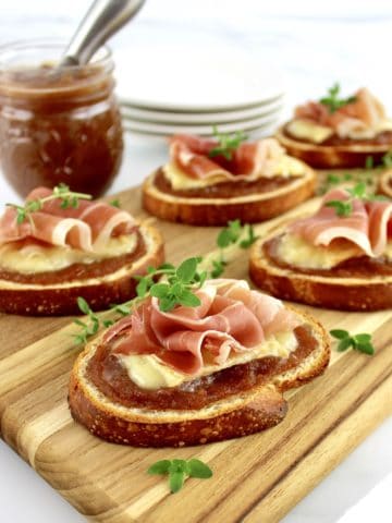Baked Brie and Prosciutto Crostini on cutting board with apple butter jar and round white plates in background