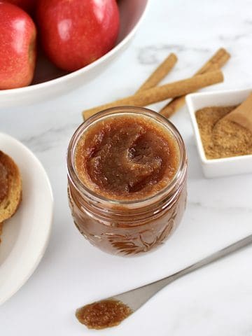 Slow Cooker Apple Butter in open glass jar with apples in background with cinnamon sticks and toast