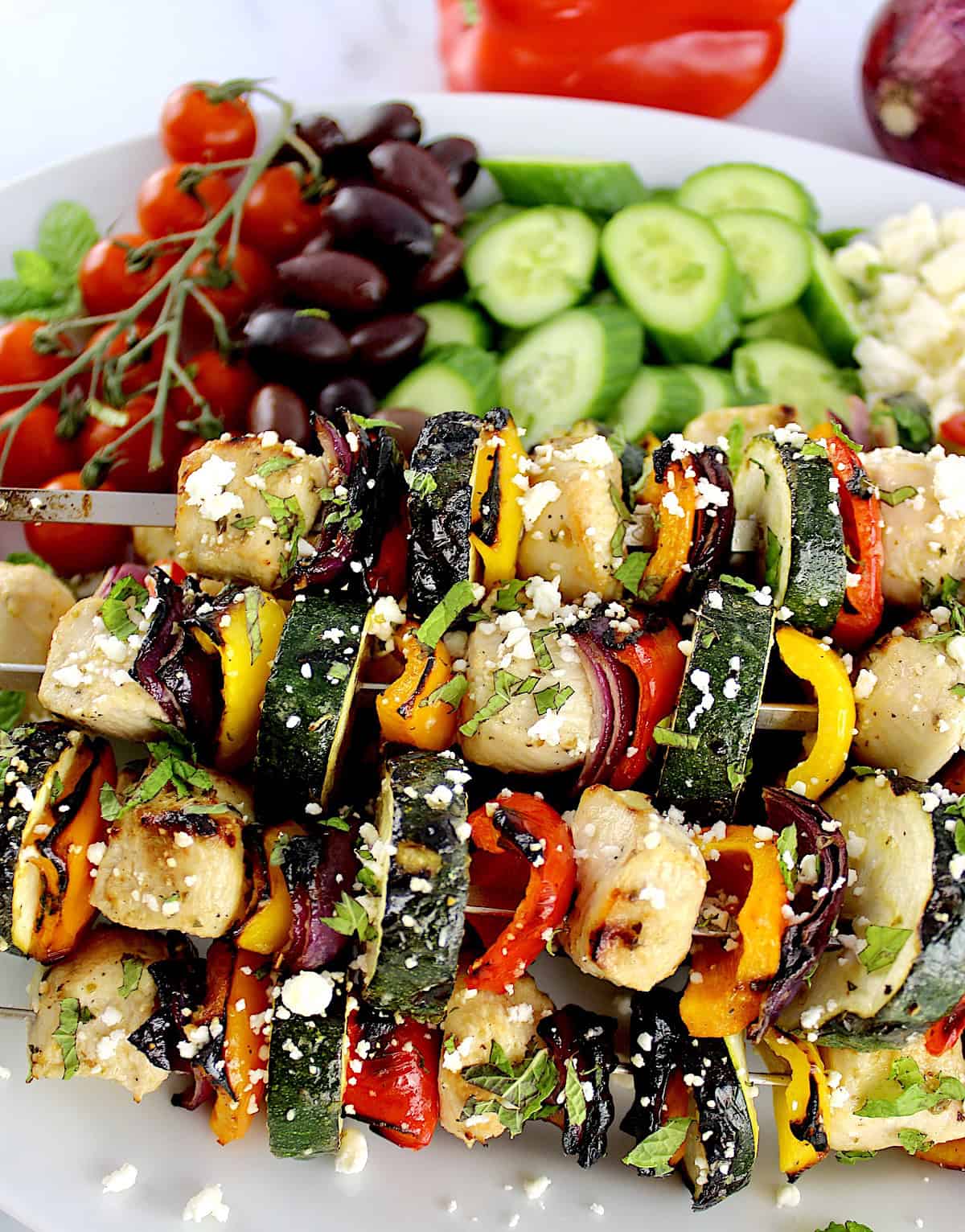 closeup of Greek Chicken Kebabs with cucumbers, tomatoes and olives in background