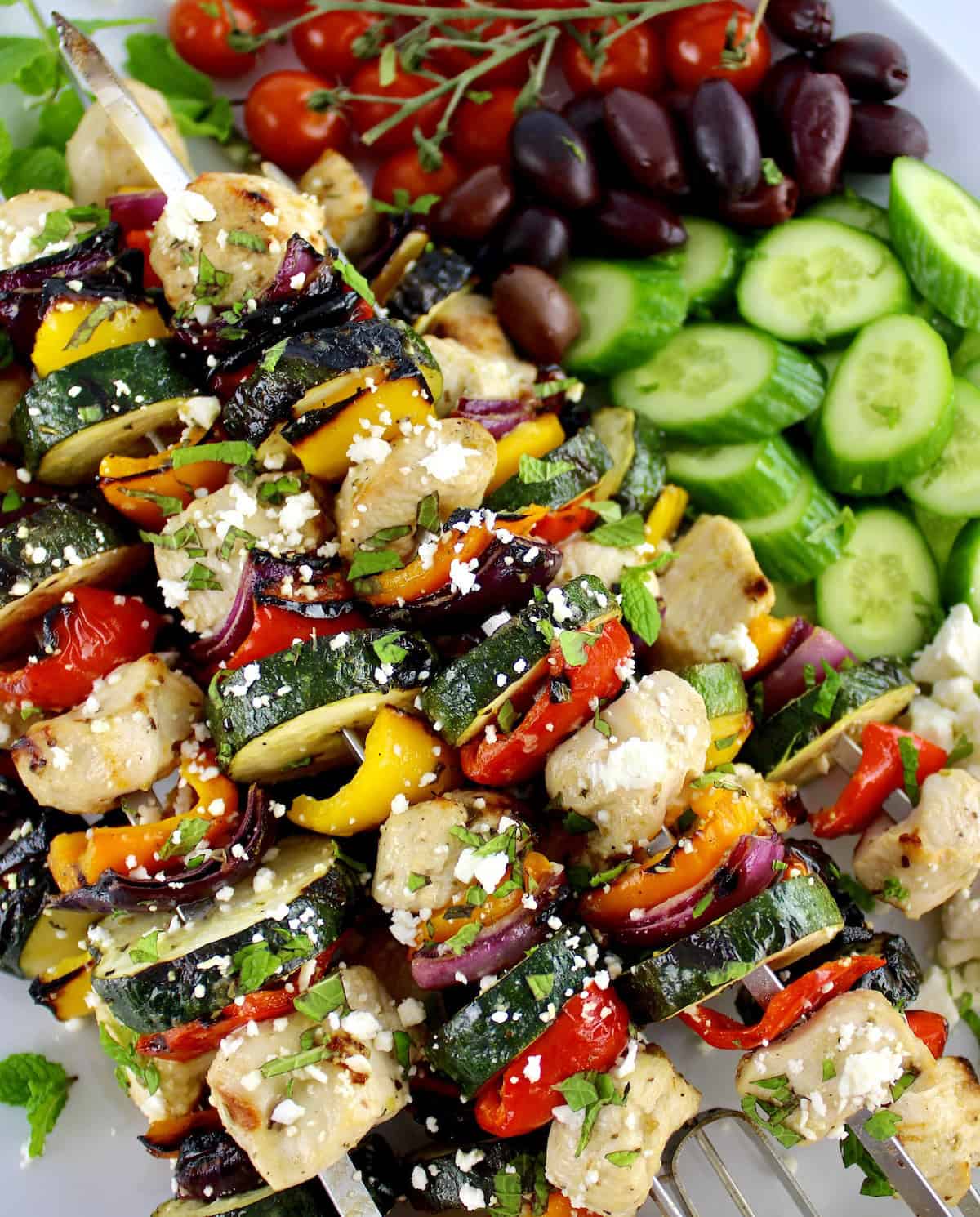 closeup of Greek Chicken Kebabs with cucumbers, tomatoes and olives in background