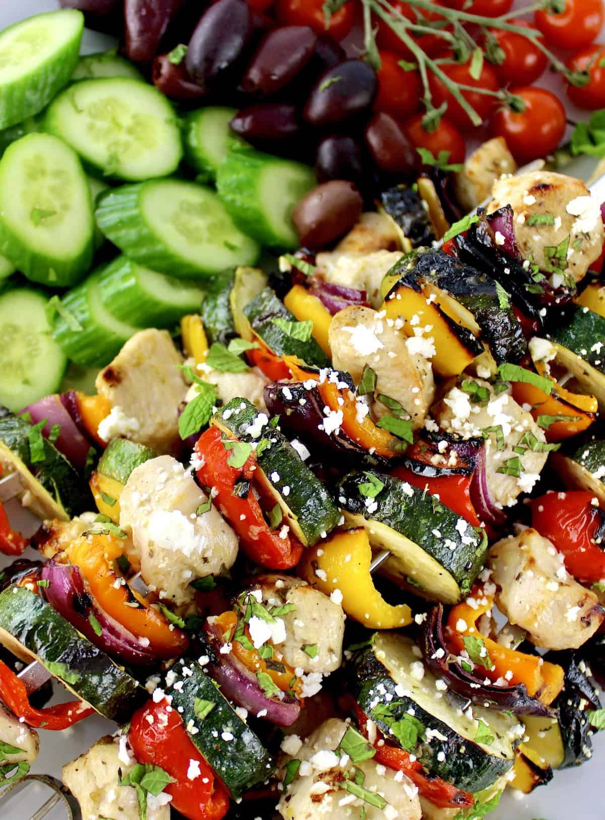 closeup of Greek Chicken Kebabs with cucumbers, tomatoes and olives in background