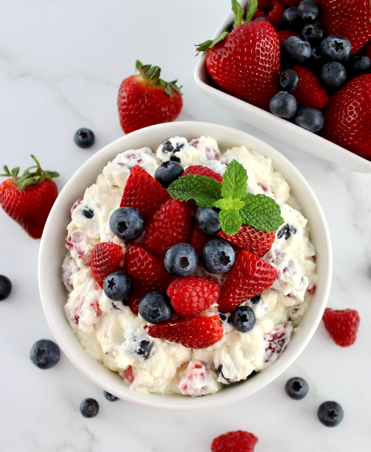 Berry Cheesecake Salad in white bowl with fresh berries and mint on top with berries in background