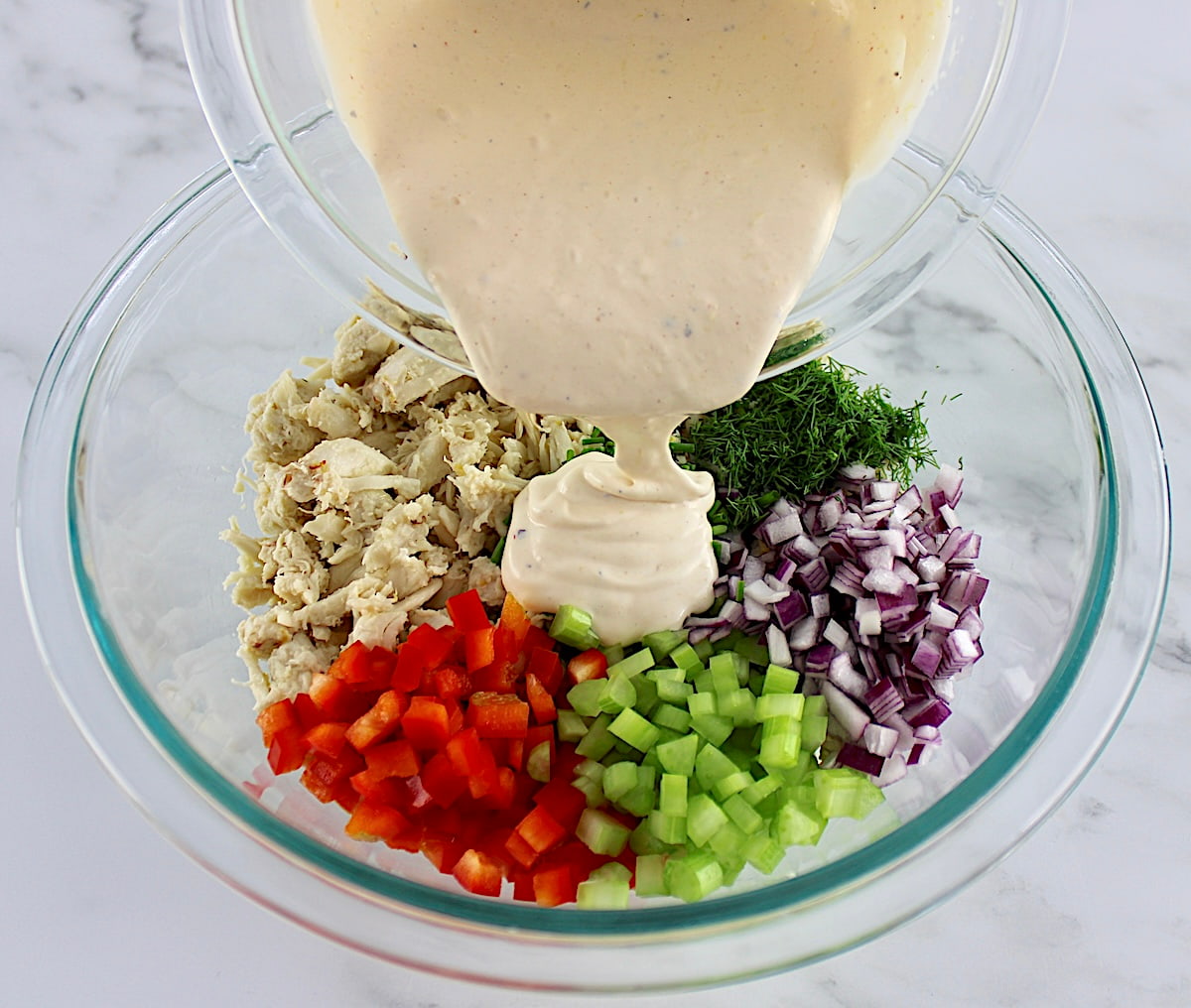 crab meat, herbs, red onion, celery and red bell pepper all chopped in glass bowl in sections with dressing pouring over the top