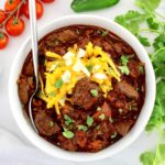 Slow Cooker Texas Chili in white bowl with spoon and shredded cheddar, diced onions and cilantro on top