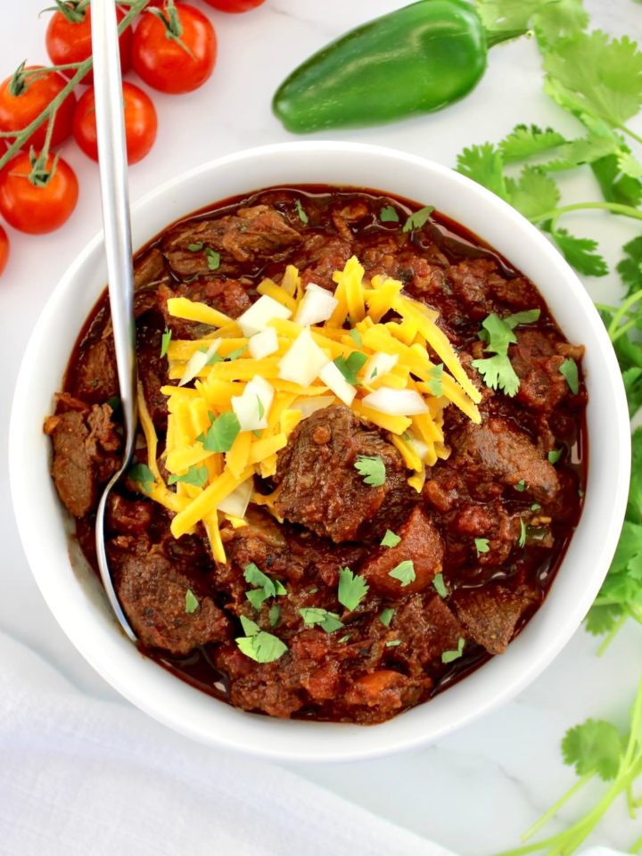 Slow Cooker Texas Chili in white bowl with spoon and shredded cheddar, diced onions and cilantro on top