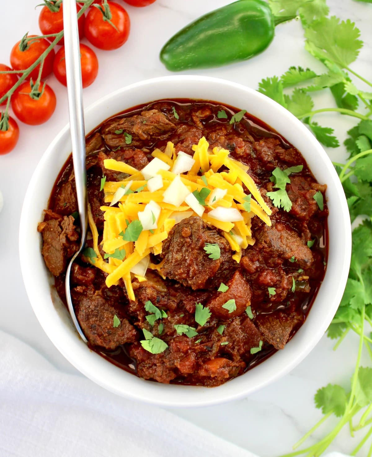Slow Cooker Texas Chili in white bowl with spoon and shredded cheddar, diced onions and cilantro on top