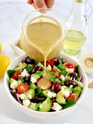 Greek Salad Dressing being poured over salad in white bowl