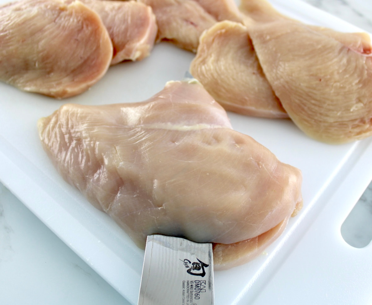 raw chicken breast being sliced in half on white cutting board