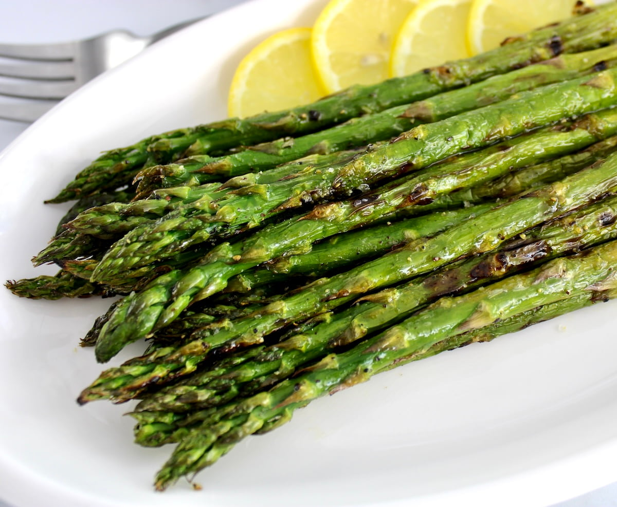closeup of Easy Grilled Asparagus on white plate with lemon slices in back