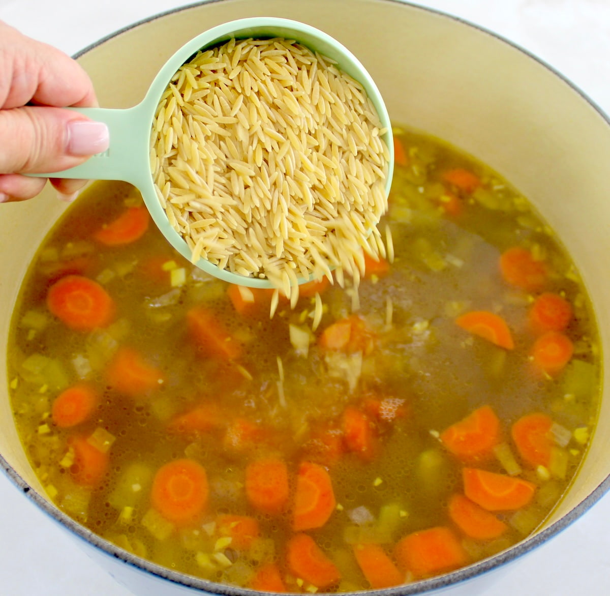 orzo being poured into chicken soup in dutch oven