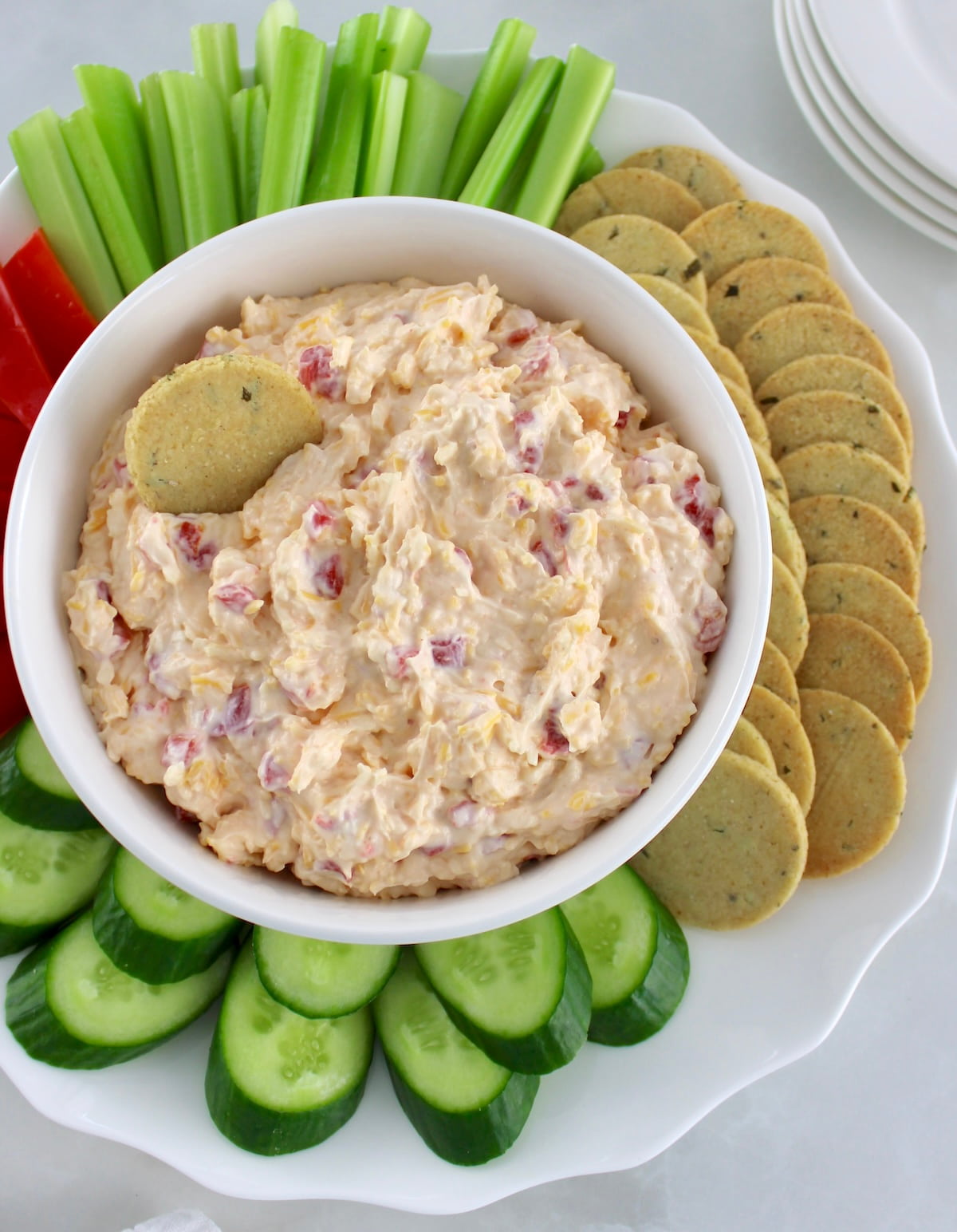 overhead view of Pimento Cheese Spread on white plate with veggies and crackers around with one cracker in dip