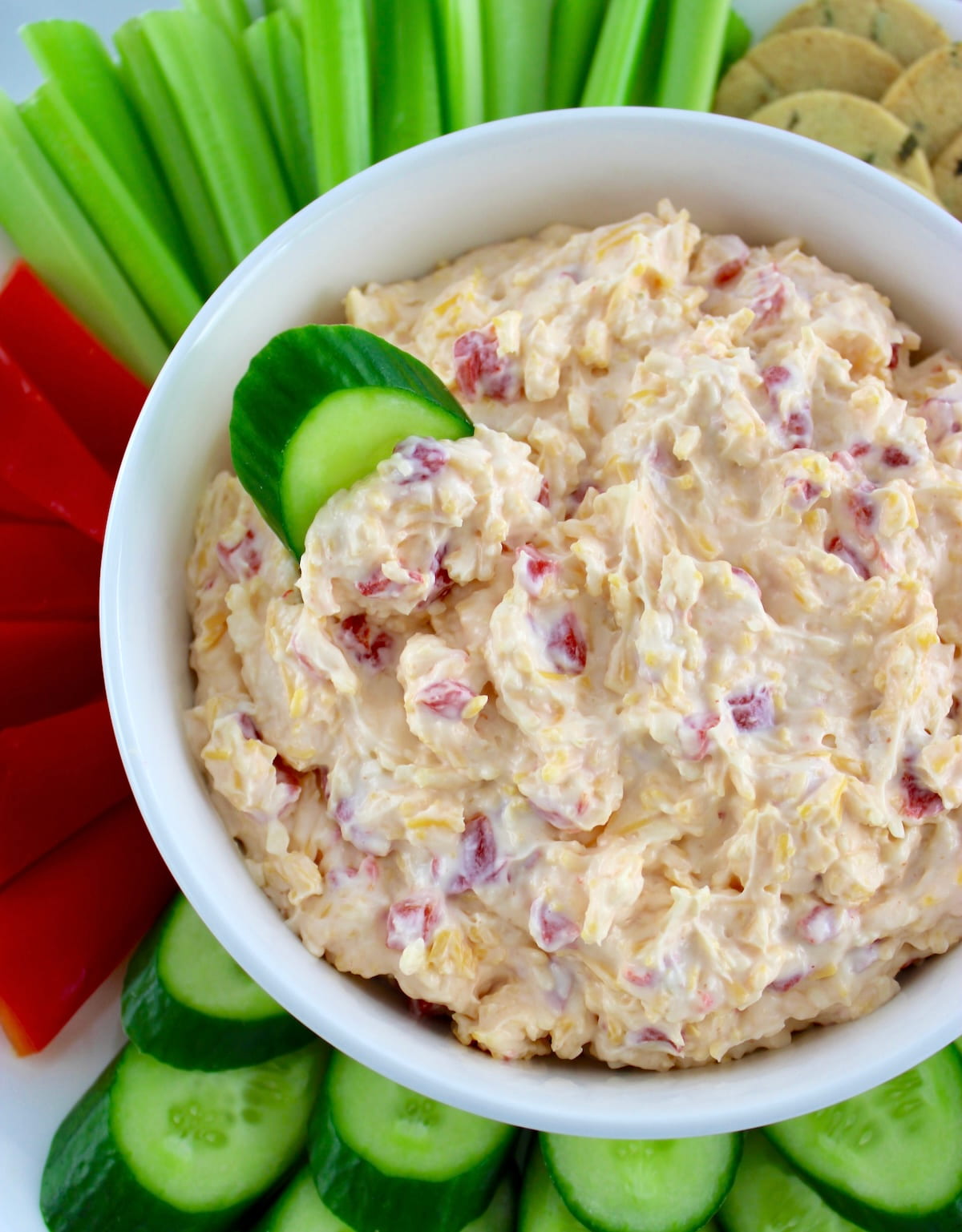 overhead view of Pimento Cheese Spread in white bowl with veggies around and cucumber slice in dip