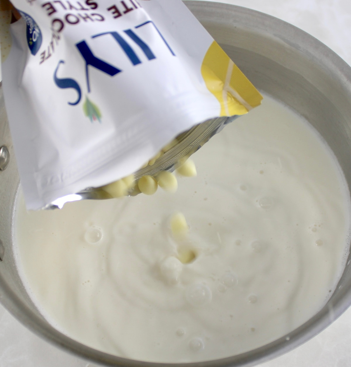 Lily's white chocolate chips being poured into hot heavy cream in pot