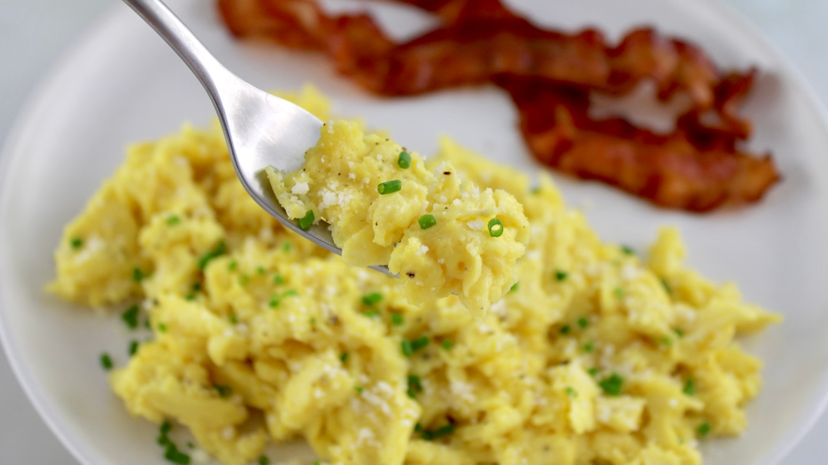 Ricotta Scrambled Eggs on white plate with bacon and fork holding up some