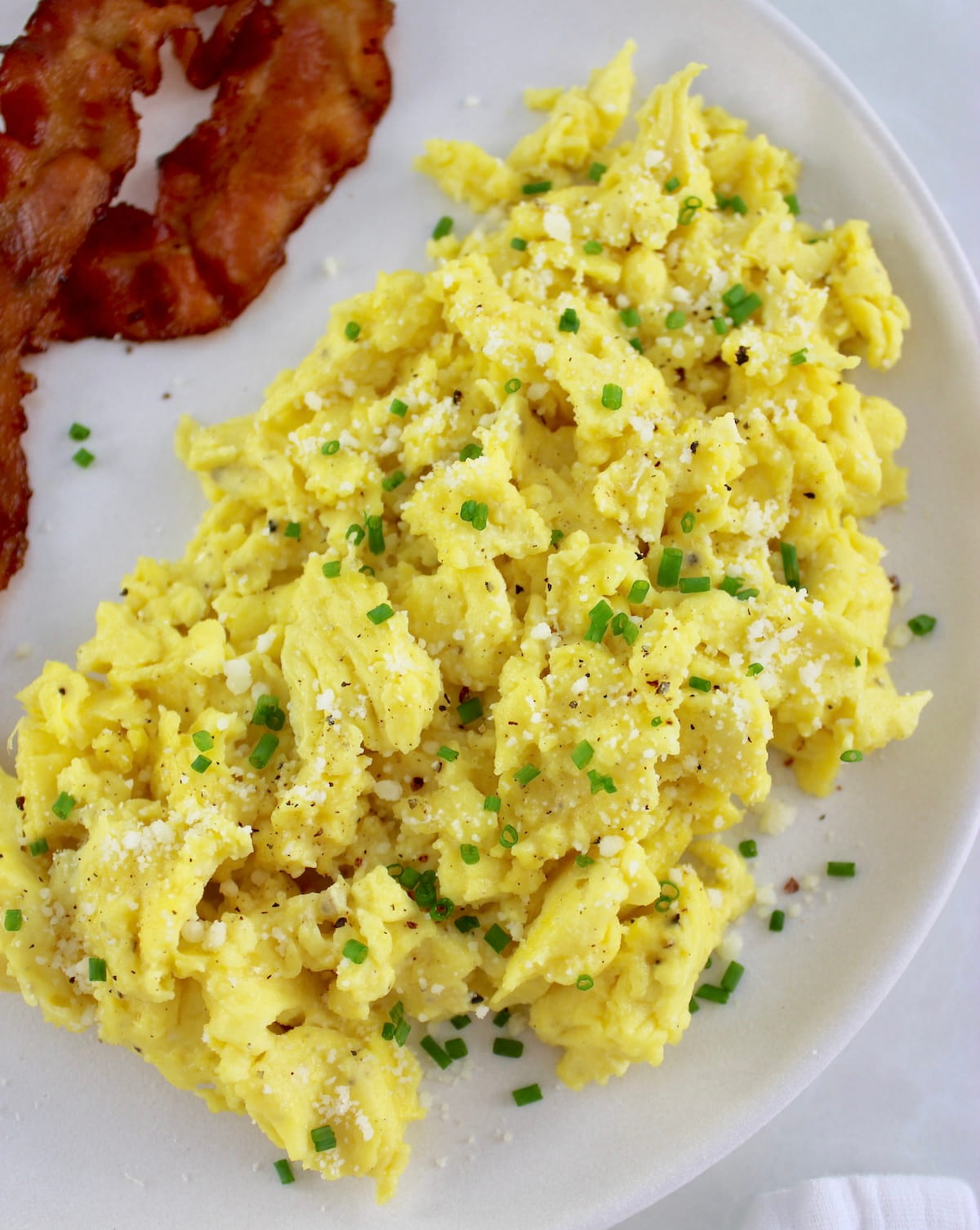 closeup of Ricotta Scrambled Eggs on white plate with bacon on side