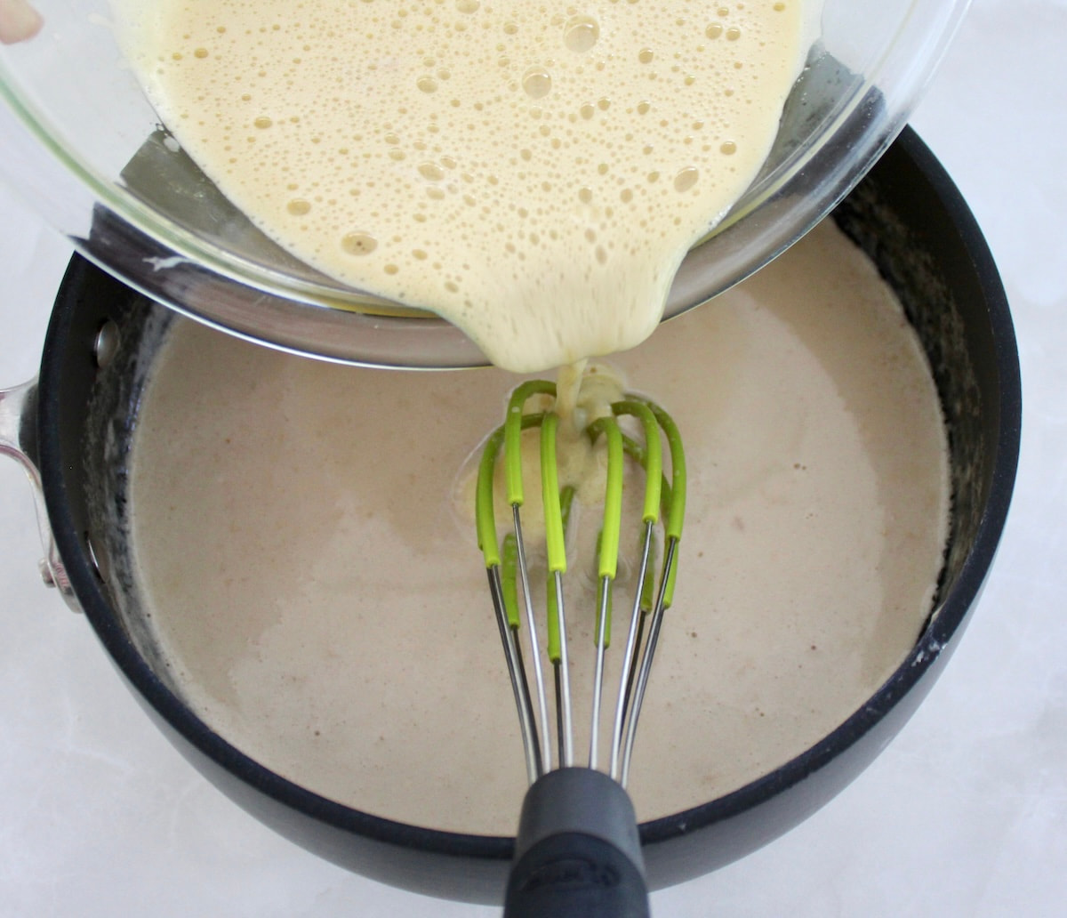tempered eggs being ladled into saucepan with hot cream and whisk