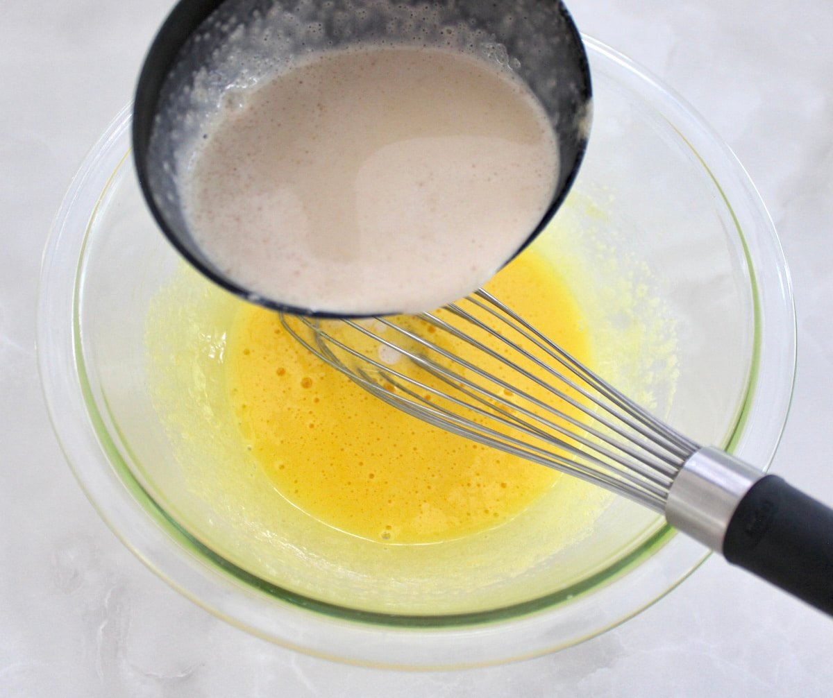 hot cream being ladled into glass bowl with eggs and whisk