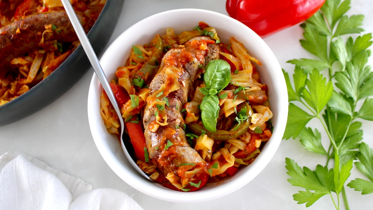 overhead view of Italian Sausage and Peppers Cabbage Skillet in white bowl with spoon and basil garnish