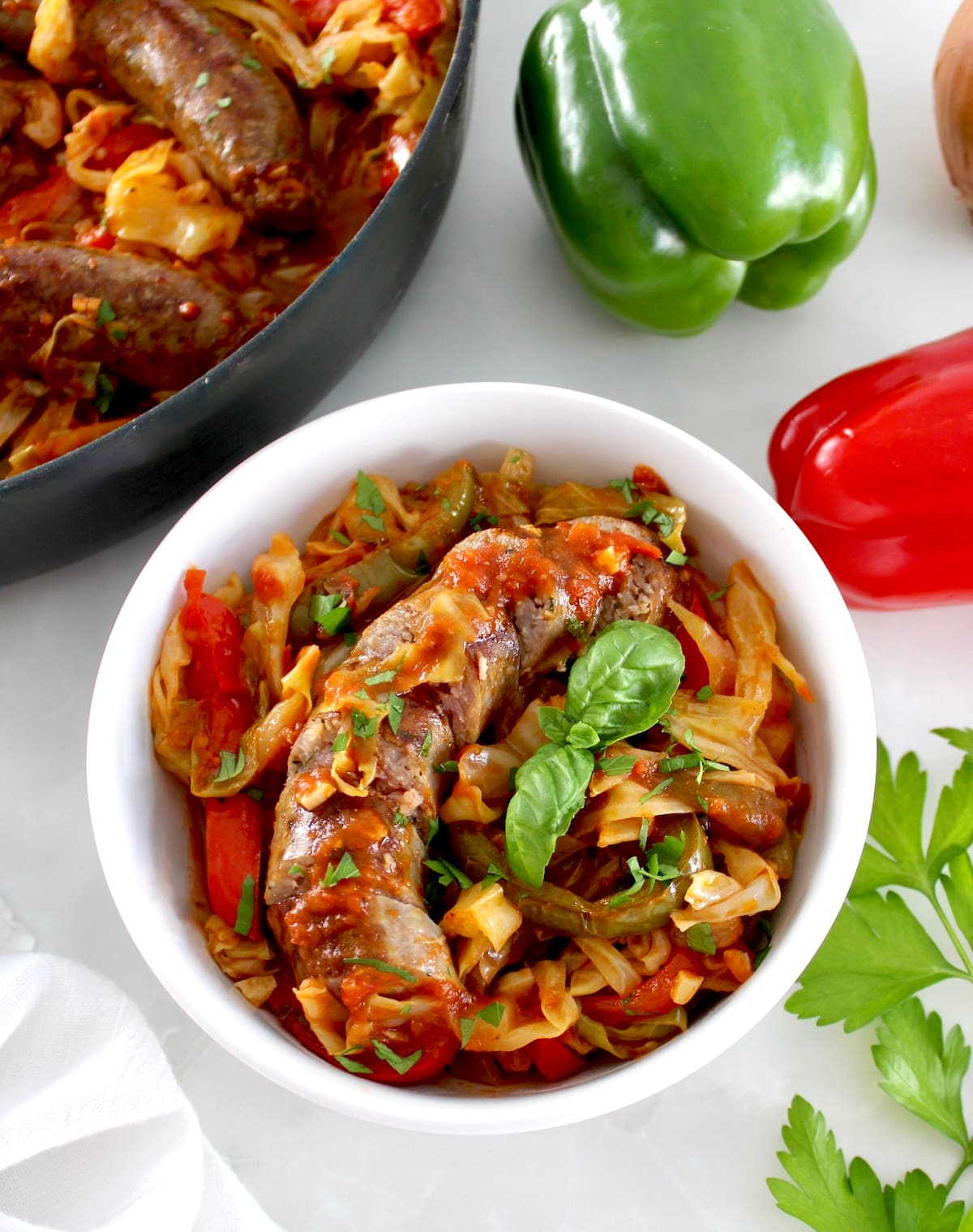 overhead view of Italian Sausage and Peppers Cabbage Skillet in white bowl with basil leaf and skillet in back