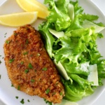 Pork Milanese on white plate with greens and shaved parmesan with 2 lemon wedges