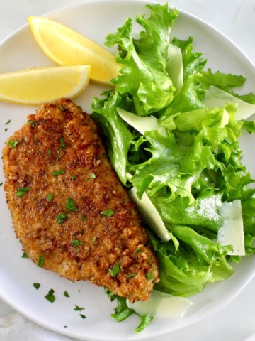Pork Milanese on white plate with greens and shaved parmesan with 2 lemon wedges