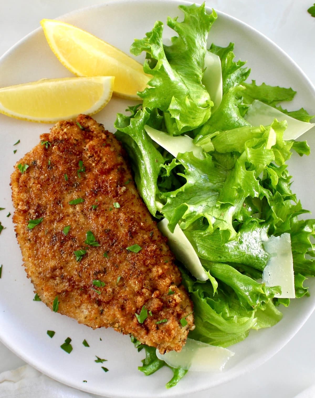 Pork Milanese on white plate with greens and shaved parmesan with 2 lemon wedges