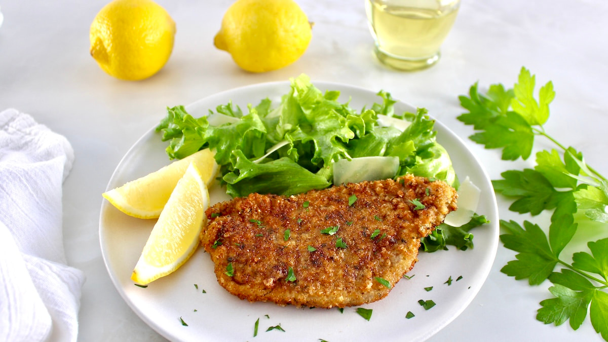 Pork Milanese on white plate with greens and 2 lemon wedges on side
