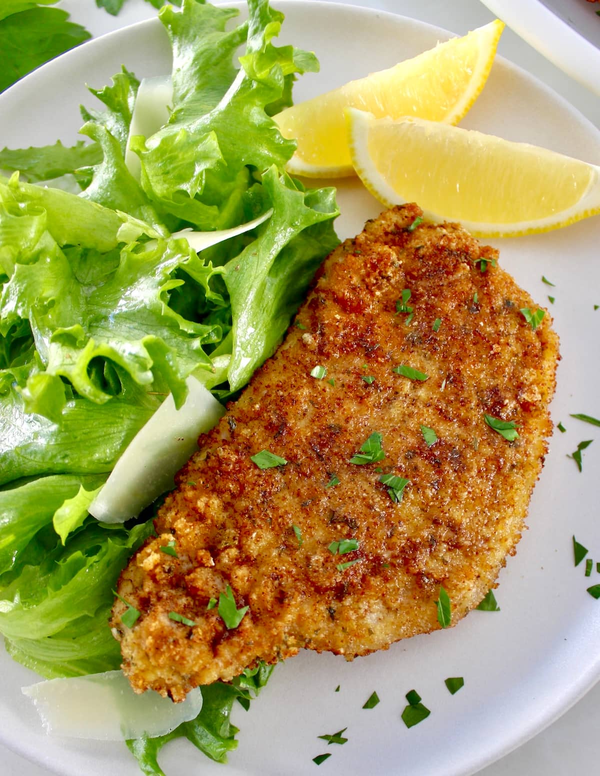 Pork Milanese on white plate with greens and shaved parmesan with 2 lemon wedges