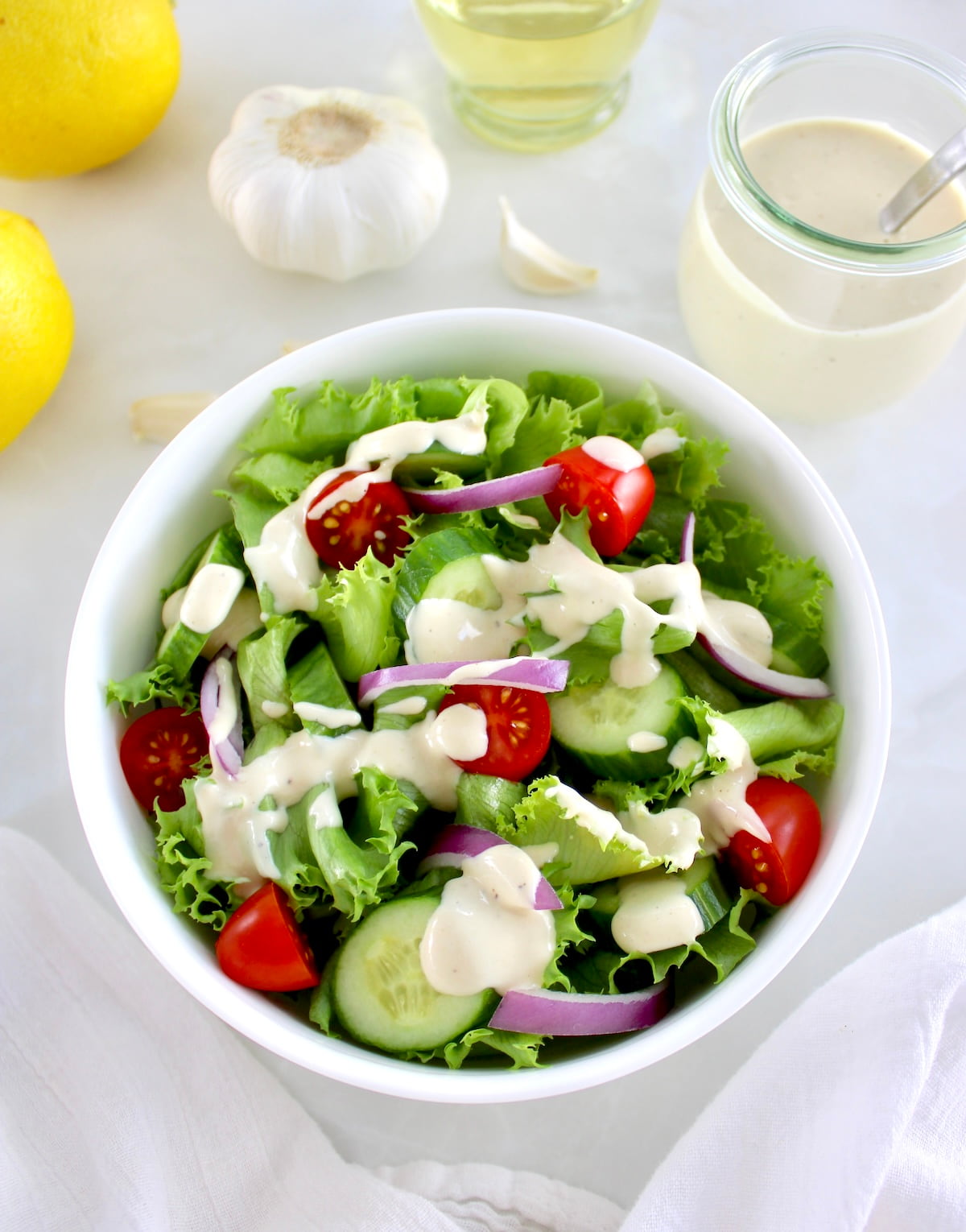 Lemon Tahini Dressing over salad in white bowl with garlic, lemon and more dressing in back