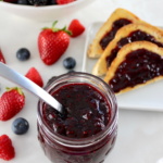 Mixed Berry Jam in open glass jar with spoon, berries and toasted in back