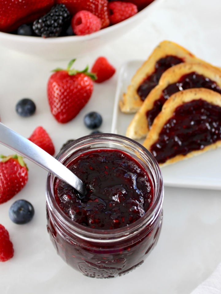 Mixed Berry Jam in open glass jar with spoon, berries and toasted in back