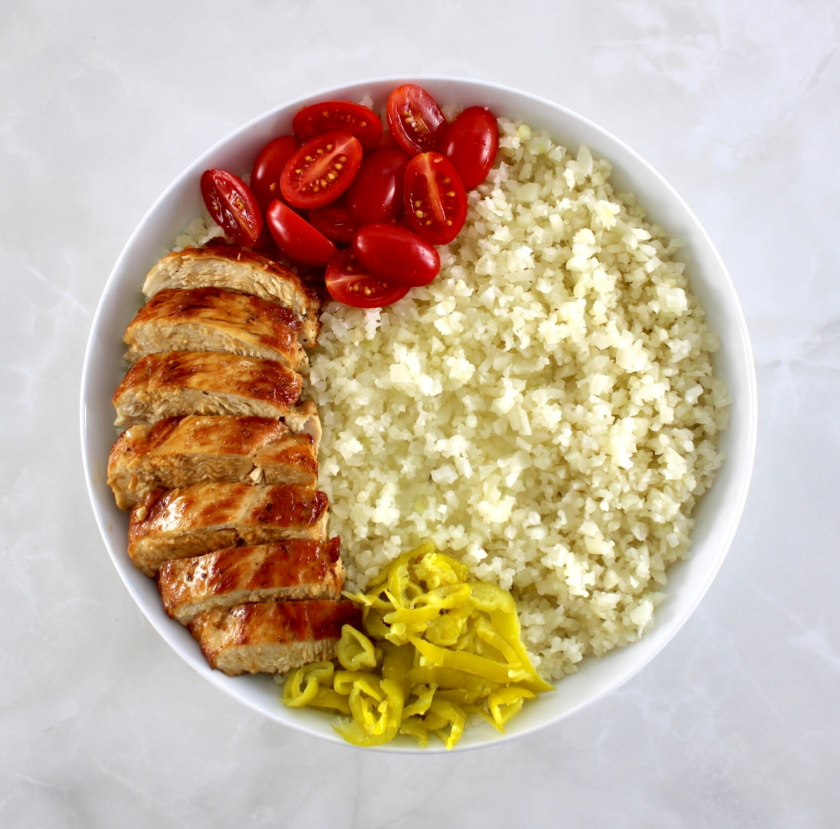 white bowl with cauliflower rice, sliced chicken, pepperoncini and halved cherry tomatoes