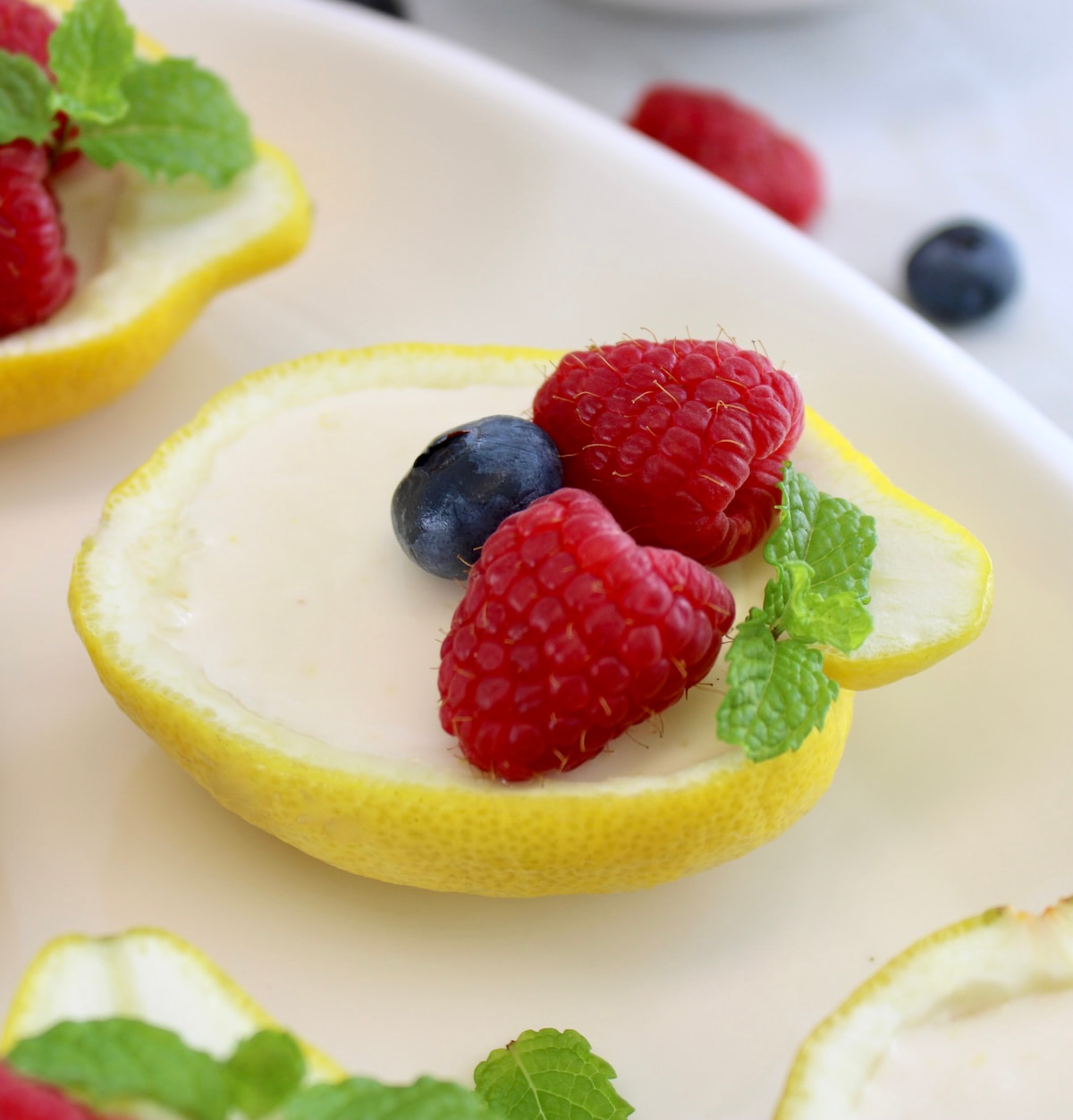 closeup of Lemon Posset in lemon half on white plate with fresh berries and mint on top