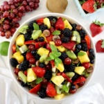 Easy Fruit Salad in white bowl with fresh mint on top with grapes and strawberries in back.