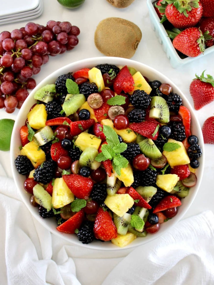 Easy Fruit Salad in white bowl with fresh mint on top with grapes and strawberries in back.