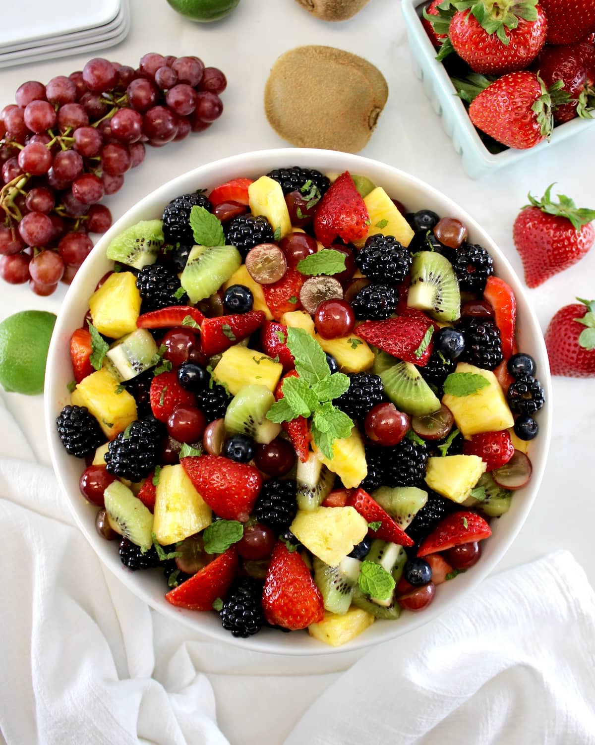 Easy Fruit Salad in white bowl with fresh mint on top with grapes and strawberries in back.