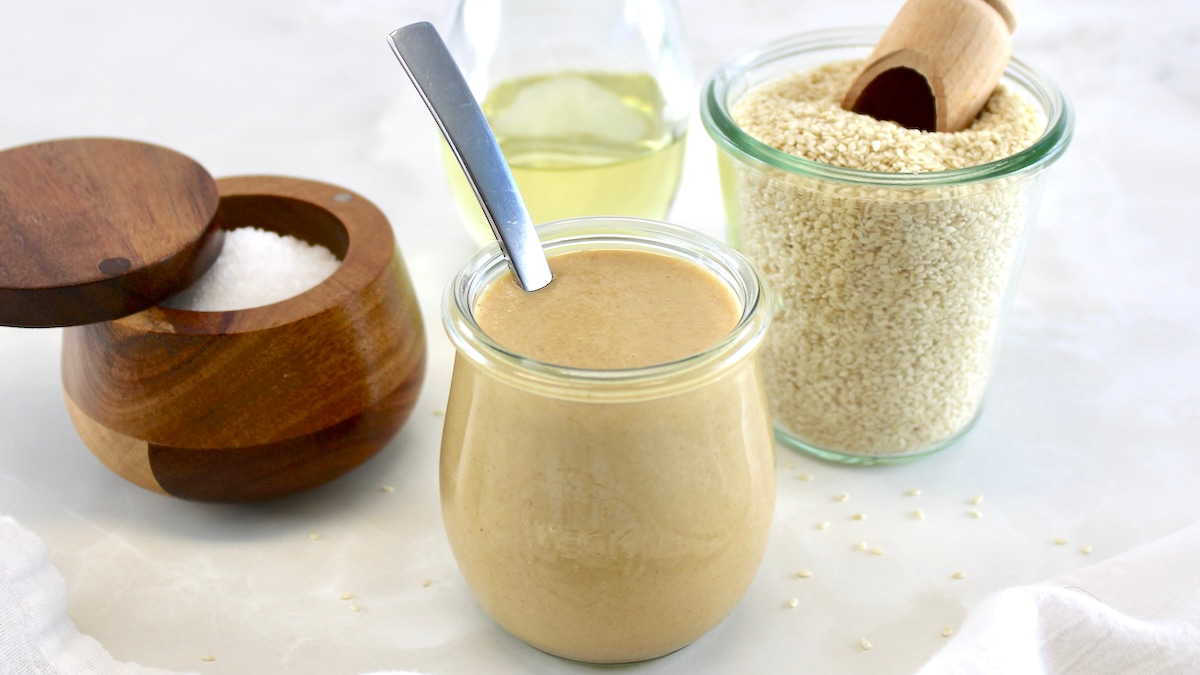 side view of Homemade Tahini in open glass jar with spoon and avocado oil, salt and sesame seeds in back