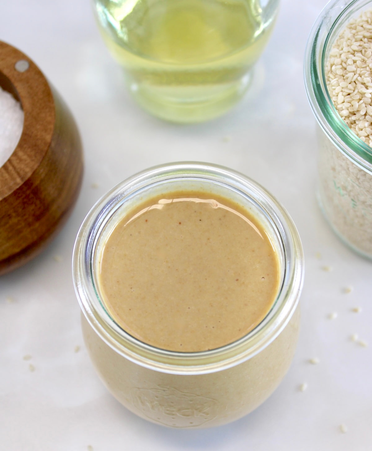 overhead view of Homemade Tahini in open glass jar with avocado oil and sesame seeds in back